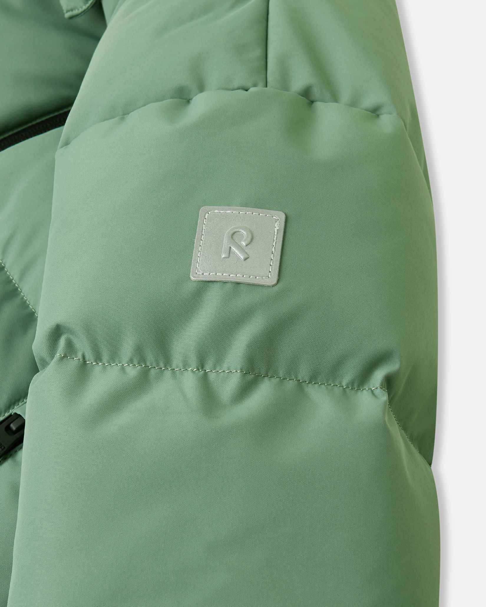 Close-up of the sleeve on the green clay Reima Paimio children's puffer coat, highlighting the square reflective Reima 'R' logo patch that enhances visibility during short winter days.