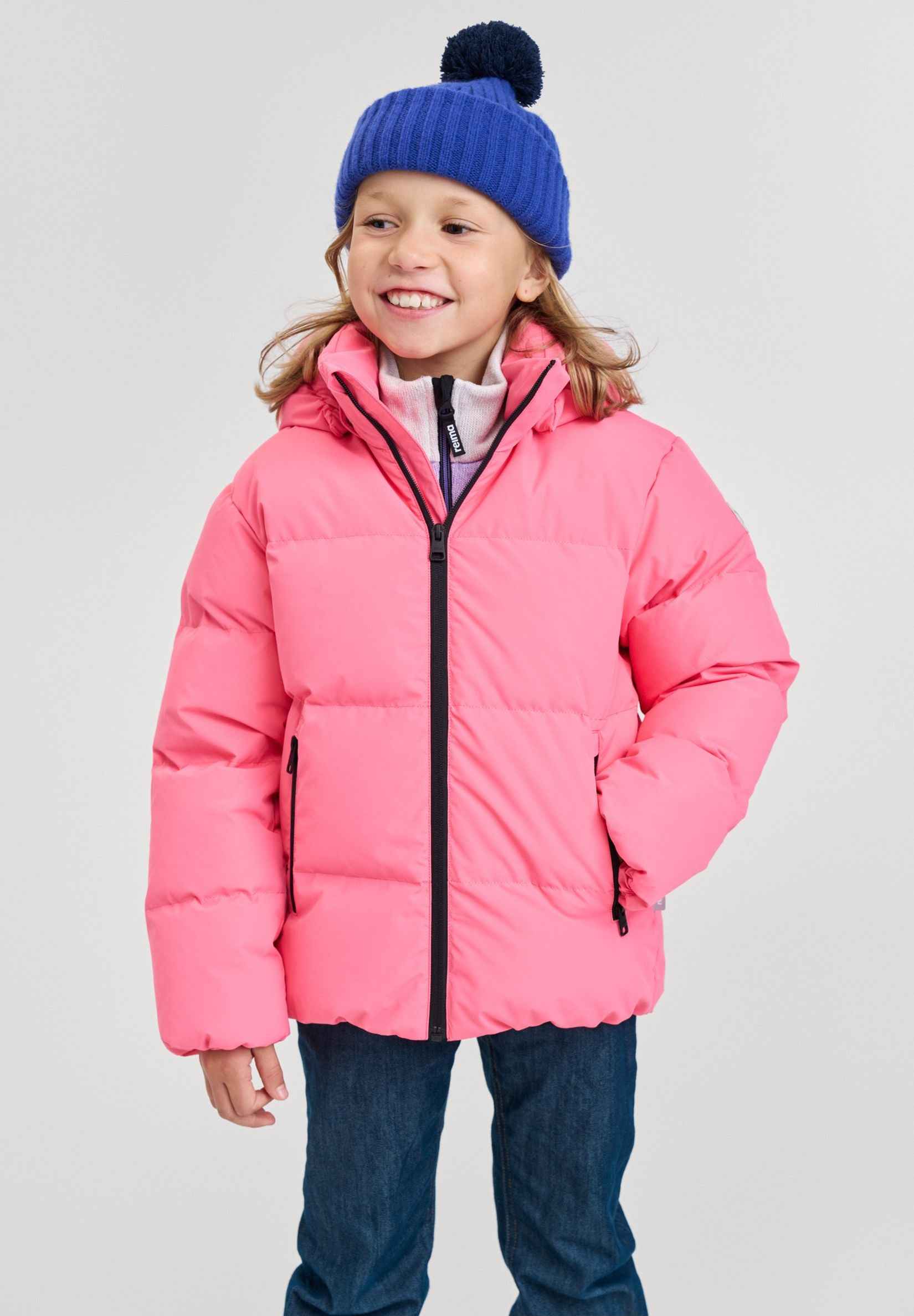 Lifestyle portrait of a young smiling girl wearing the soft coral pink Reima Paimio kids' winter down puffer jacket, paired with a blue knit beanie, showcasing its lightweight, cozy fit for active outdoor play.