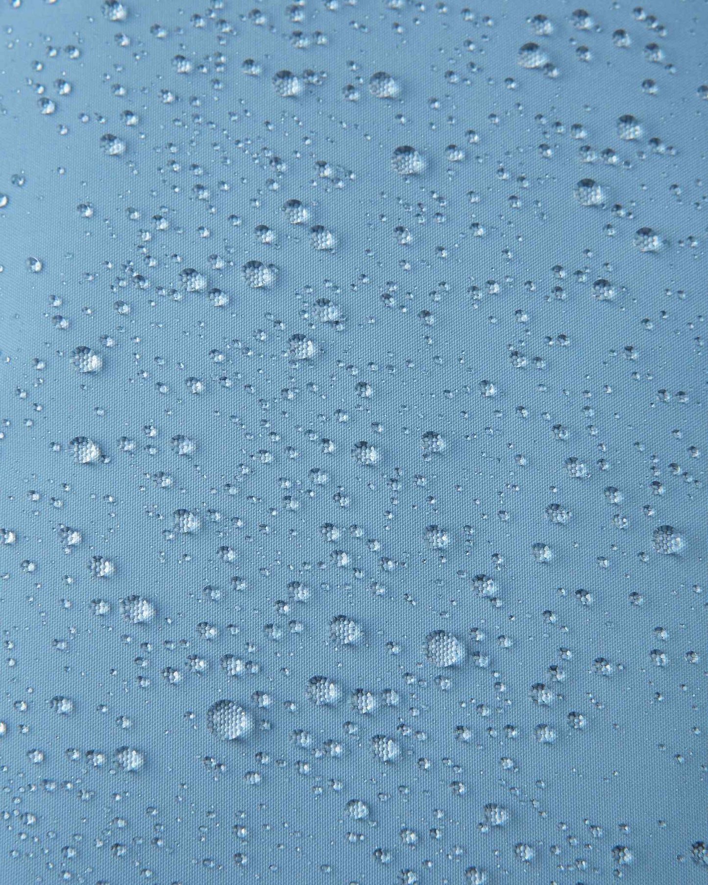 Macro close-up of water droplets beading across the surface of the cloudy blue outer shell of the Reima Paimio kids' down jacket, highlighting the water-repellent, windproof, and breathable textile finish.