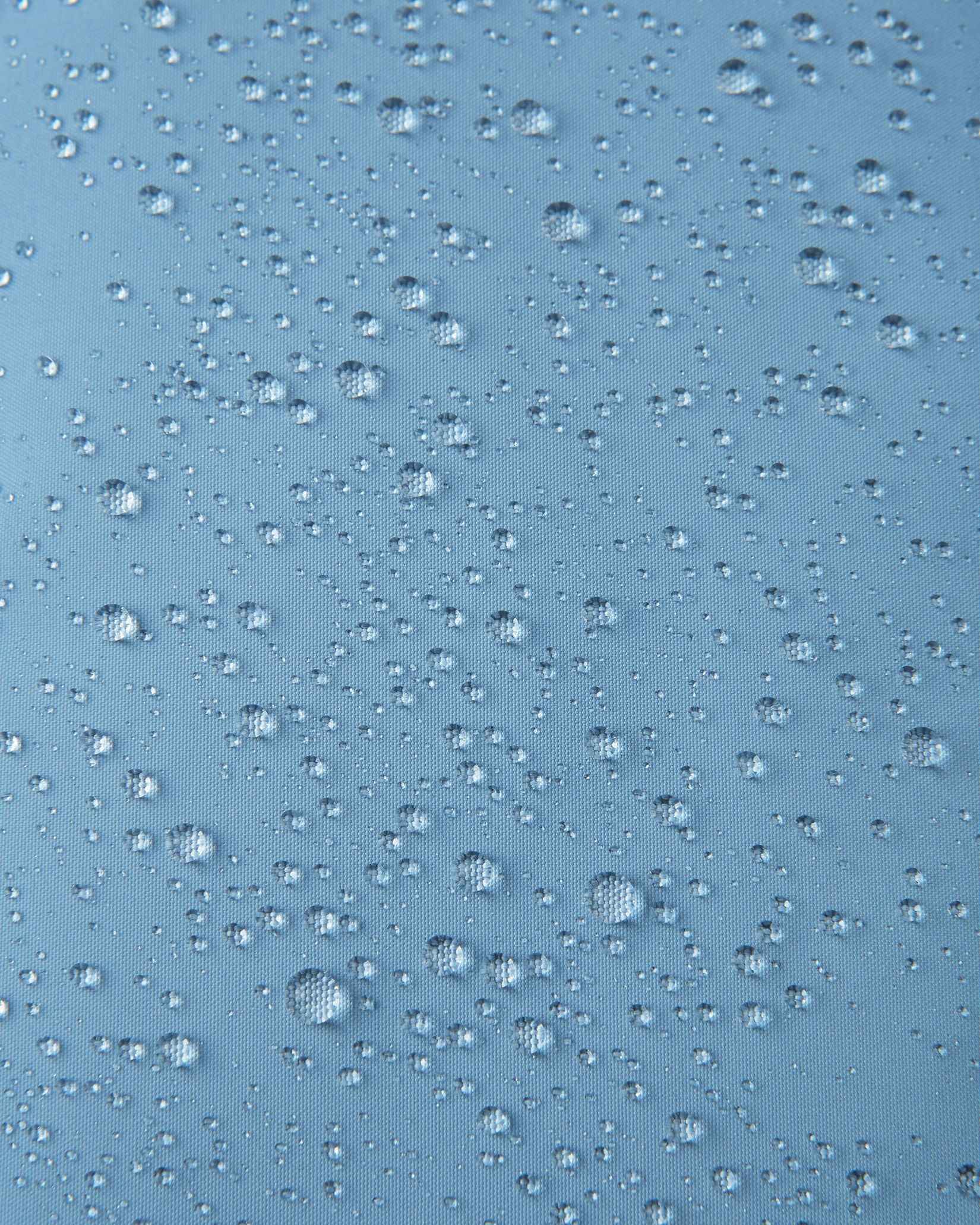 Macro close-up of water droplets beading across the surface of the cloudy blue outer shell of the Reima Paimio kids' down jacket, highlighting the water-repellent, windproof, and breathable textile finish.