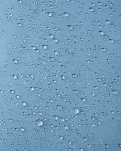 Macro close-up of water droplets beading across the surface of the cloudy blue outer shell of the Reima Paimio kids' down jacket, highlighting the water-repellent, windproof, and breathable textile finish.