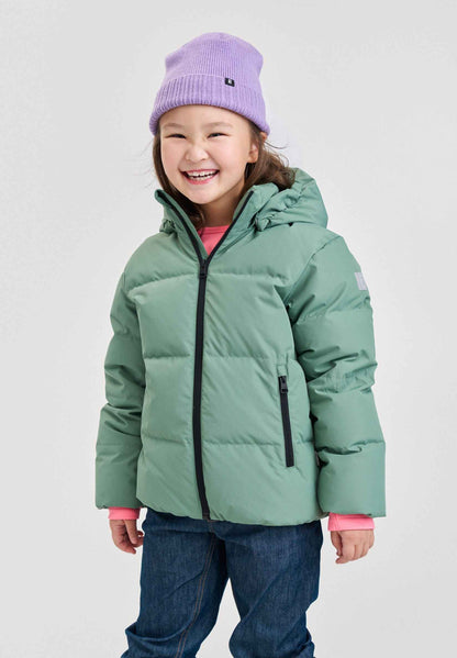 Lifestyle portrait of a young smiling girl wearing the green clay Reima Paimio kids' winter down puffer jacket, paired with a purple beanie, showcasing its lightweight and cozy fit for outdoor play.