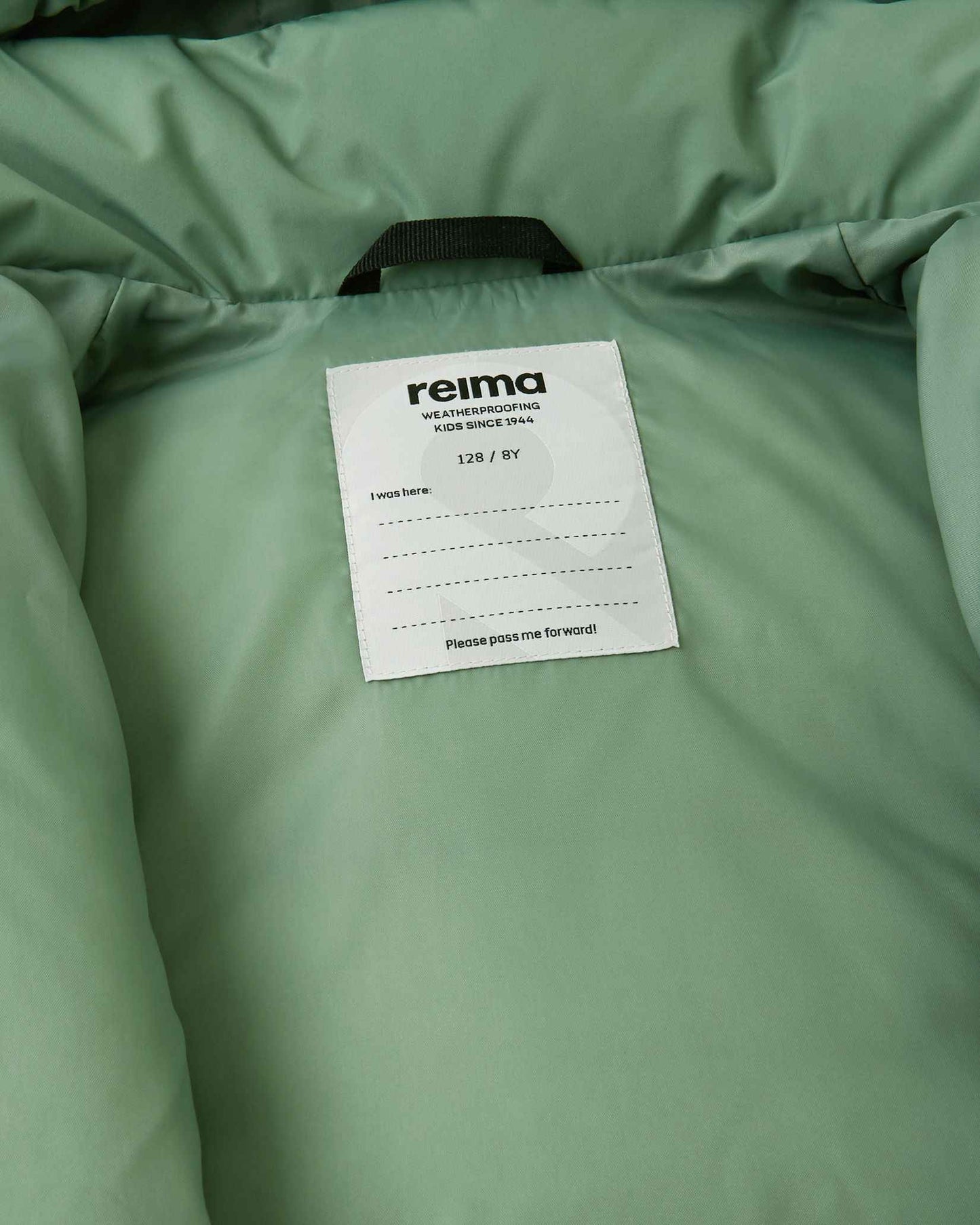 Inside view of the Reima Paimio youth winter jacket, displaying the Reima weatherproofing label, sizing information, and a customizable "I was here" name tag block for school or camp.