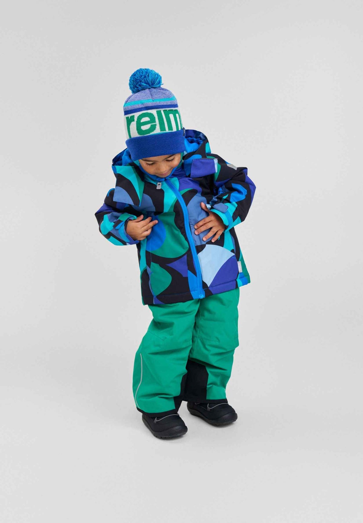 A child modeling the Aquatic Reima Palsi ski jacket featuring blue and green circular patterns, paired with bright green snow pants and a blue Reima beanie.