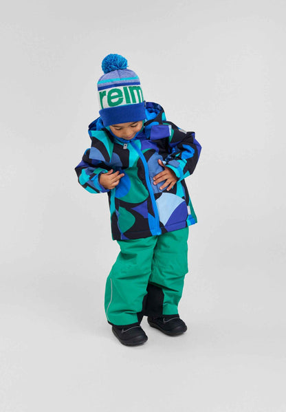 A child modeling the Aquatic Reima Palsi ski jacket featuring blue and green circular patterns, paired with bright green snow pants and a blue Reima beanie.