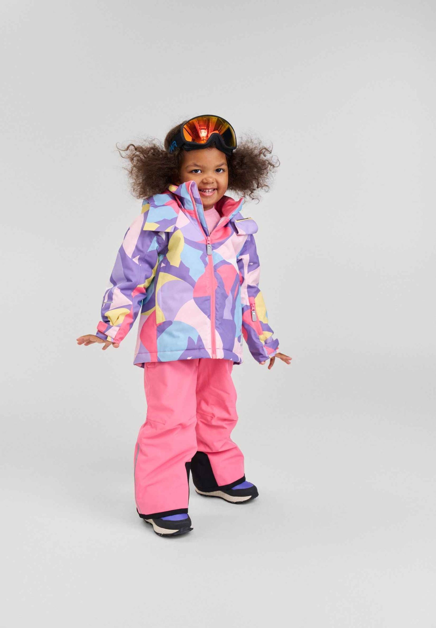A young girl stands against a light grey background wearing the Blooming Lilac Reima Palsi ski jacket. Bright pink snow pants and dark ski boots complete her outfit. She is smiling broadly with large ski goggles resting on top of her curly hair.