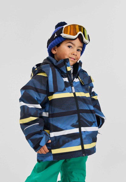 A child modeling the Blue Ocean Reima Palsi ski jacket featuring dark blue, light blue, white, and yellow geometric patterns. The jacket is paired with bright green snow Reima Proxima pants, a blue knit beanie, and reflective ski goggles.