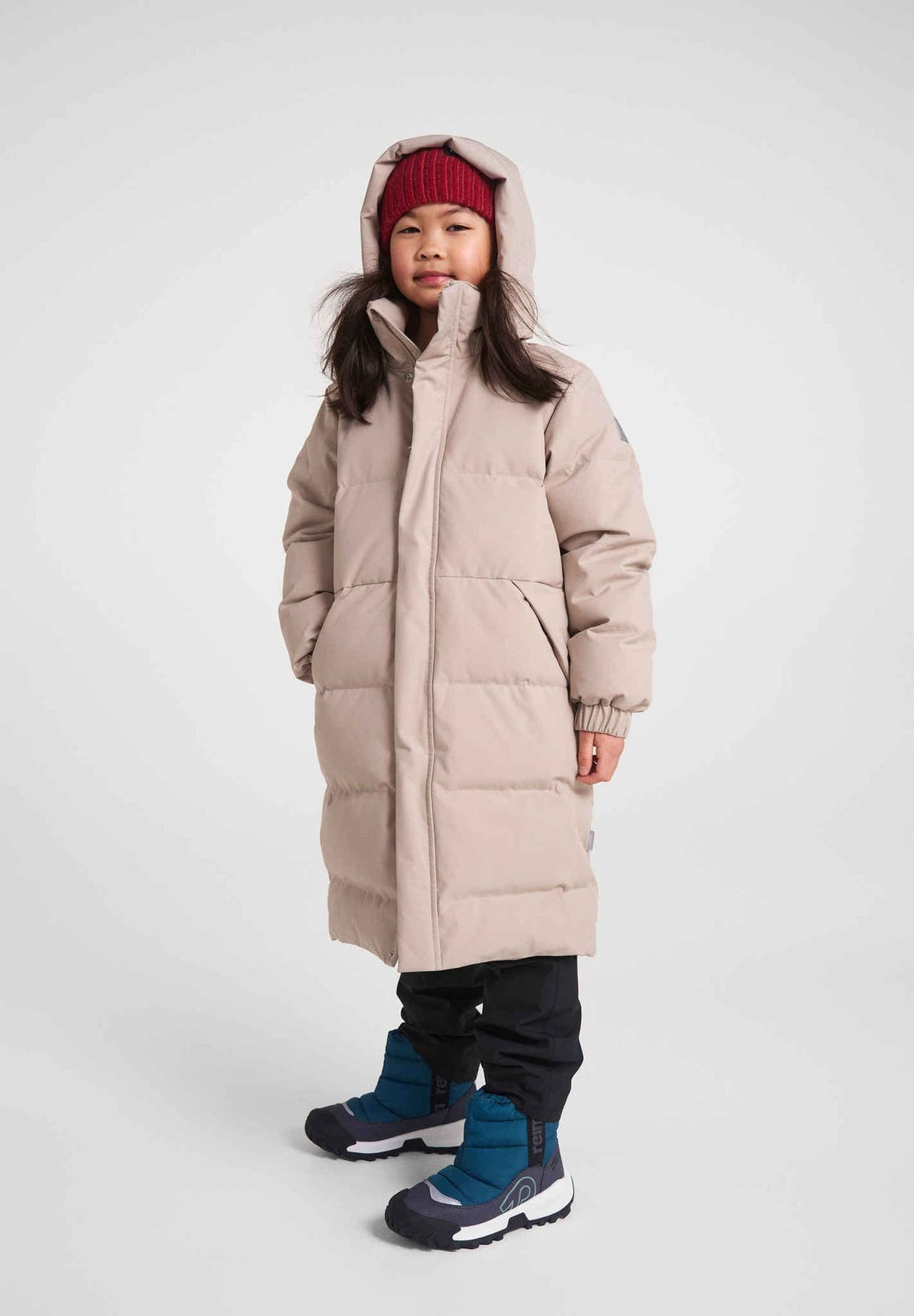 Full-length view of a young girl modeling the Reima Parkkala long down winter coat in earthy beige, paired with a red knit beanie, black snow pants, and blue winter boots against a light grey studio background.