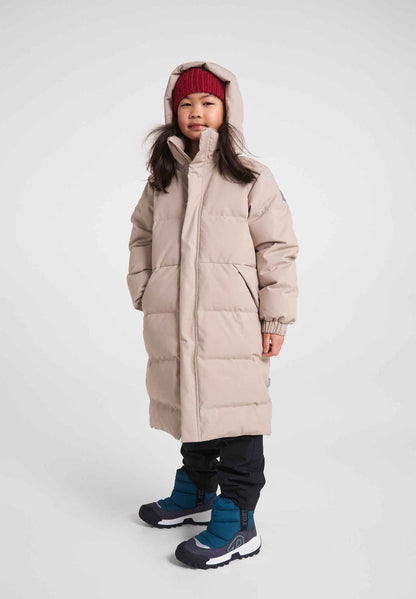 Full-length view of a young girl modeling the Reima Parkkala long down winter coat in earthy beige, paired with a red knit beanie, black snow pants, and blue winter boots against a light grey studio background.