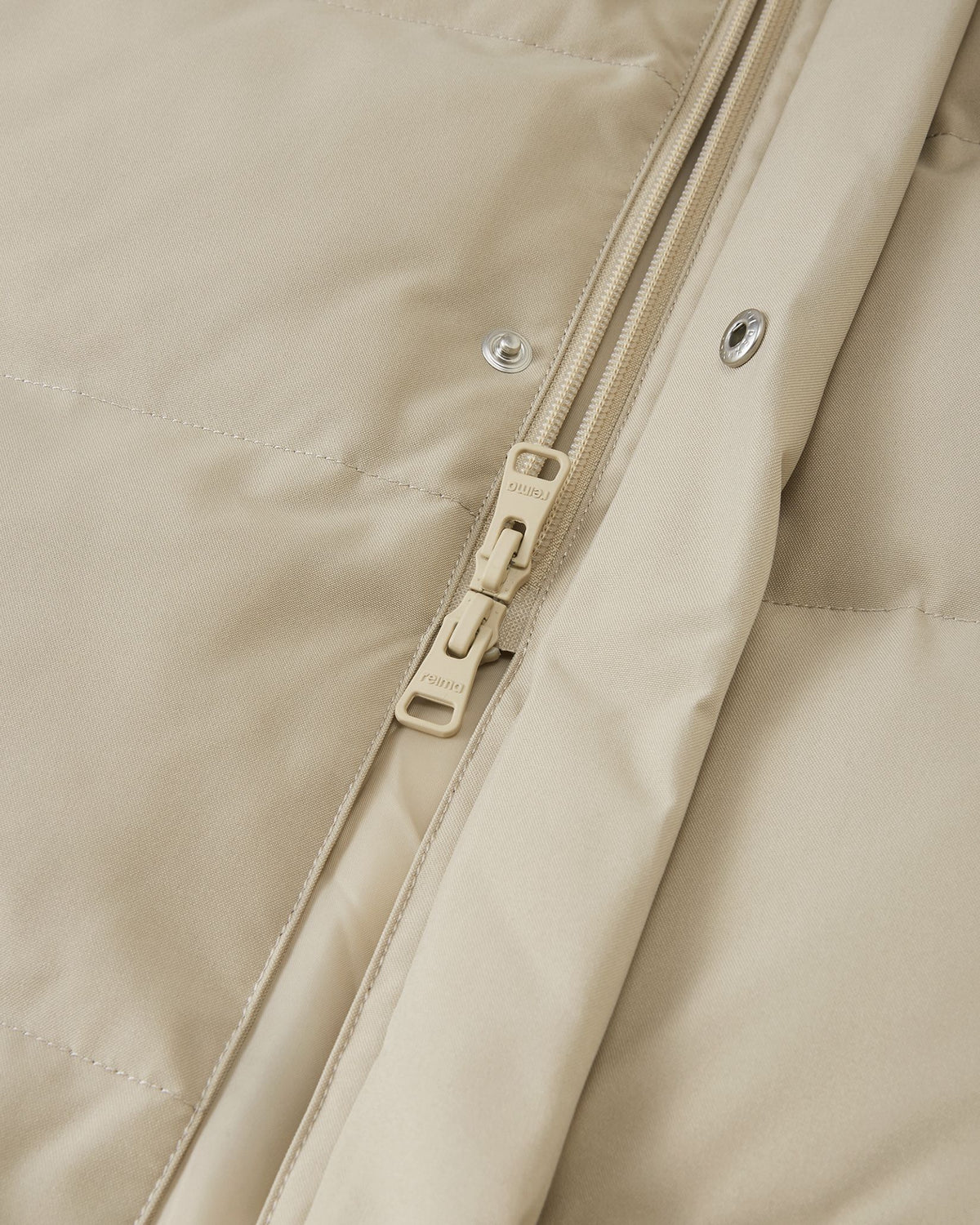 High-definition macro detail shot focusing on the central zipper of the Reima Parkkala girls' long down winter coat, showing the texture of the Earthy Beige fabric, stitching, and the specific metallic Reima branded zipper pull.