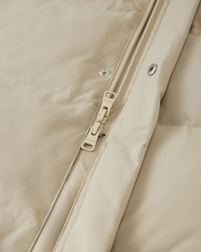 High-definition macro detail shot focusing on the central zipper of the Reima Parkkala girls' long down winter coat, showing the texture of the Earthy Beige fabric, stitching, and the specific metallic Reima branded zipper pull.