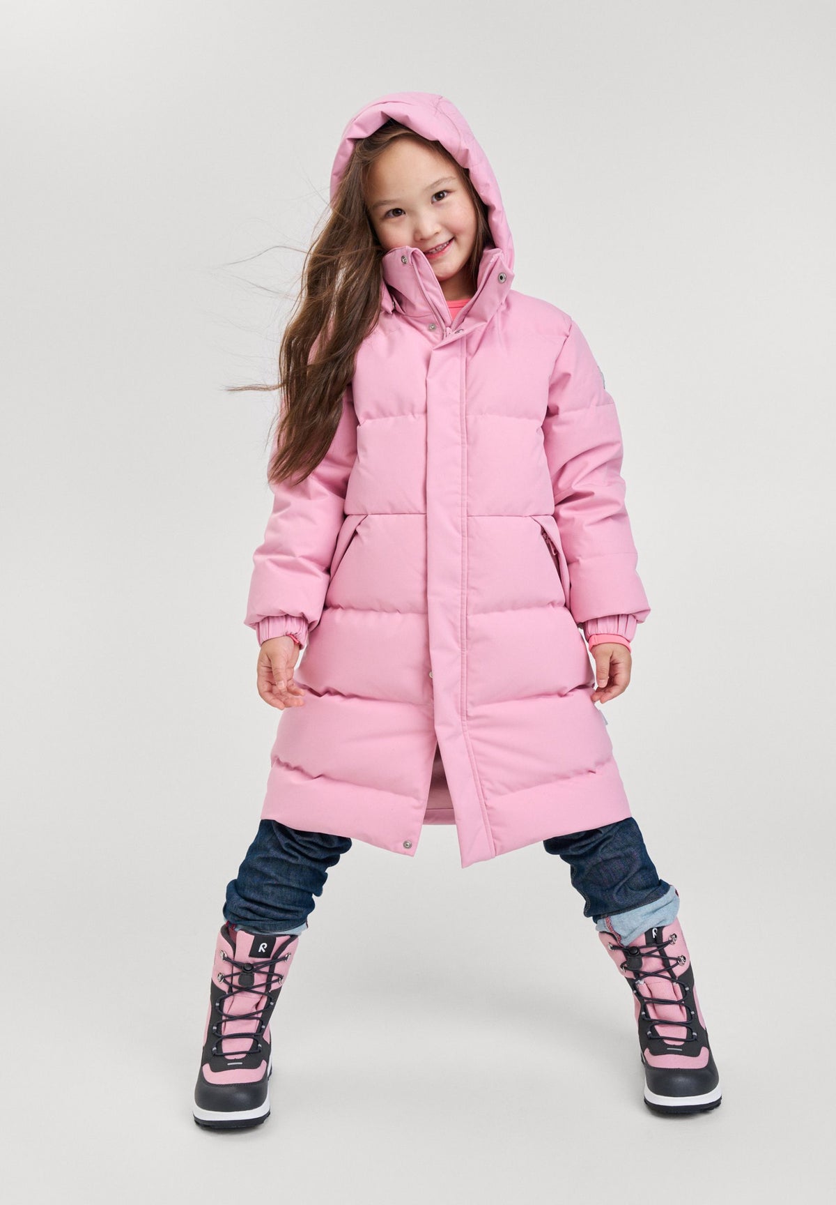 Studio lifestyle portrait of a young girl happily modeling the grey pink Reima Parkkala girls' long down puffer jacket, paired with jeans and winter boots, demonstrating the coat's stylish knee-length cut and cozy active fit.