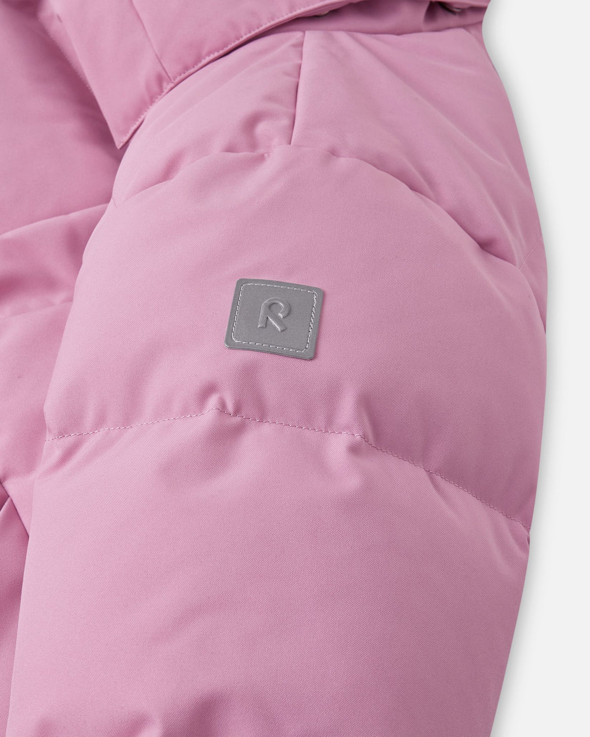 Close-up of the quilted puffy sleeve on the grey pink Reima Parkkala youth longline jacket, featuring the square high-visibility reflective Reima 'R' logo patch for enhanced safety during dark winter afternoons.