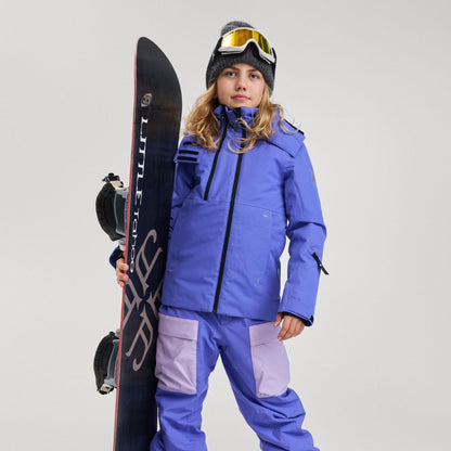 A young girl holding a snowboard while modeling the Reima Perille girls' ski jacket in breezy violet, featuring contrasting black front and chest zippers, an insulated protective hood, and designed for warm, waterproof performance on the slopes.