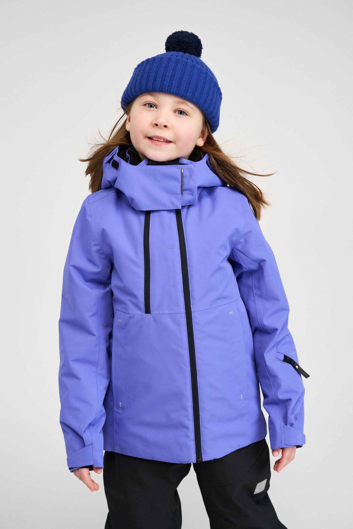 Lifestyle portrait of a young girl wearing the breezy violet Reima Perille kids' ski jacket with a blue knit beanie, showcasing the relaxed fit and warm 140g Fellex recycled insulation during winter play.