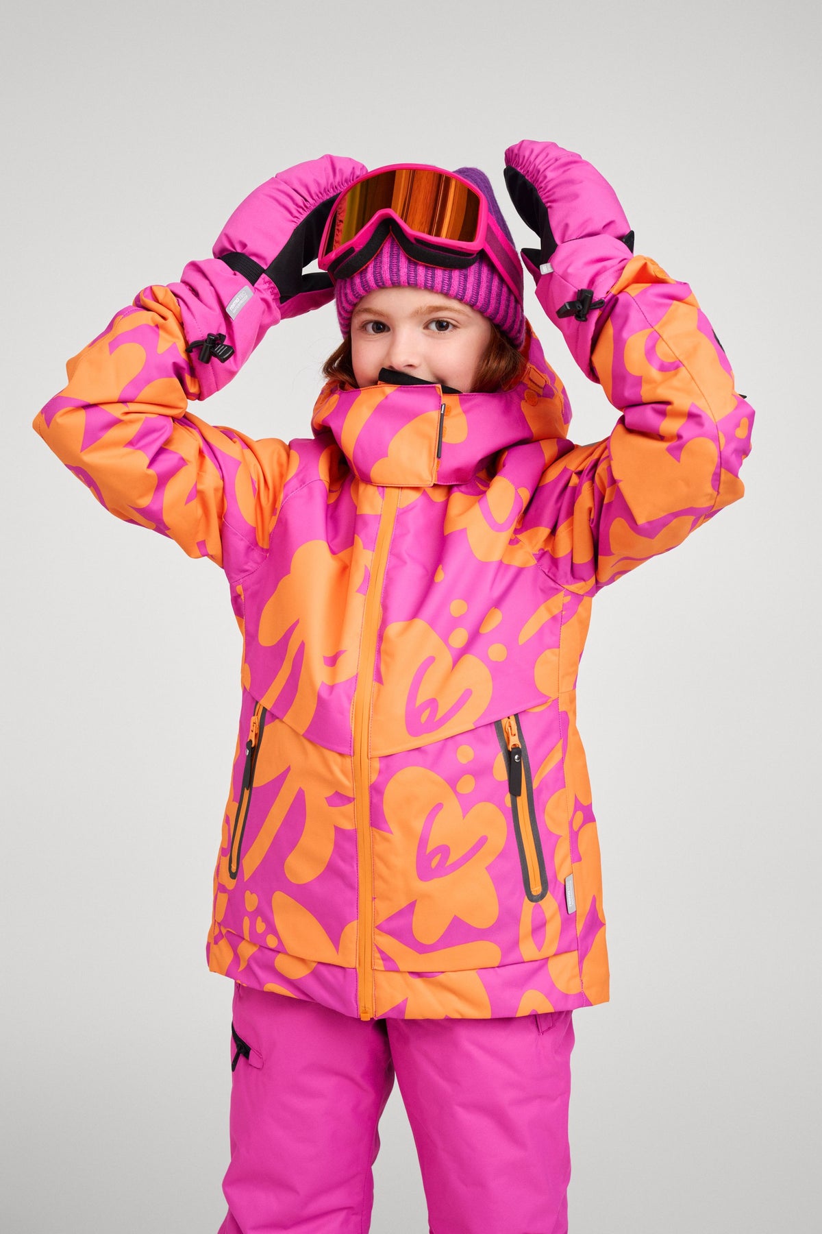 A young girl modeling the Reima Posio waterproof ski jacket in fuchsia and orange floral print, fully geared for winter sports with matching fuchsia snow pants, ski goggles, a knit beanie, and winter gloves.