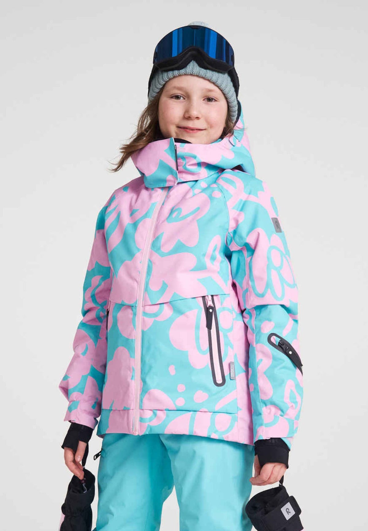 Portrait of a young girl wearing the Reima Posio high-quality girls' winter ski jacket in a light heather pink and teal floral print, styled with a grey beanie and blue ski goggles.