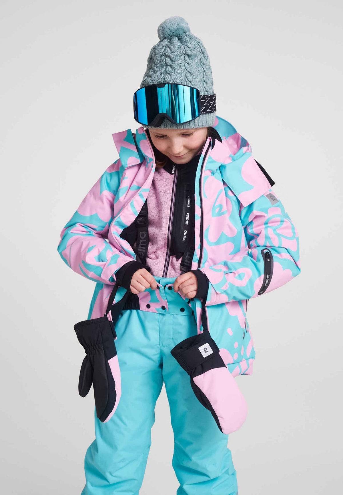 A young girl modeling a complete winter sports outfit, featuring the Reima Posio girls' ski jacket in light heather floral, paired with matching light blue snow pants, winter mittens on leashes, and a knit beanie.