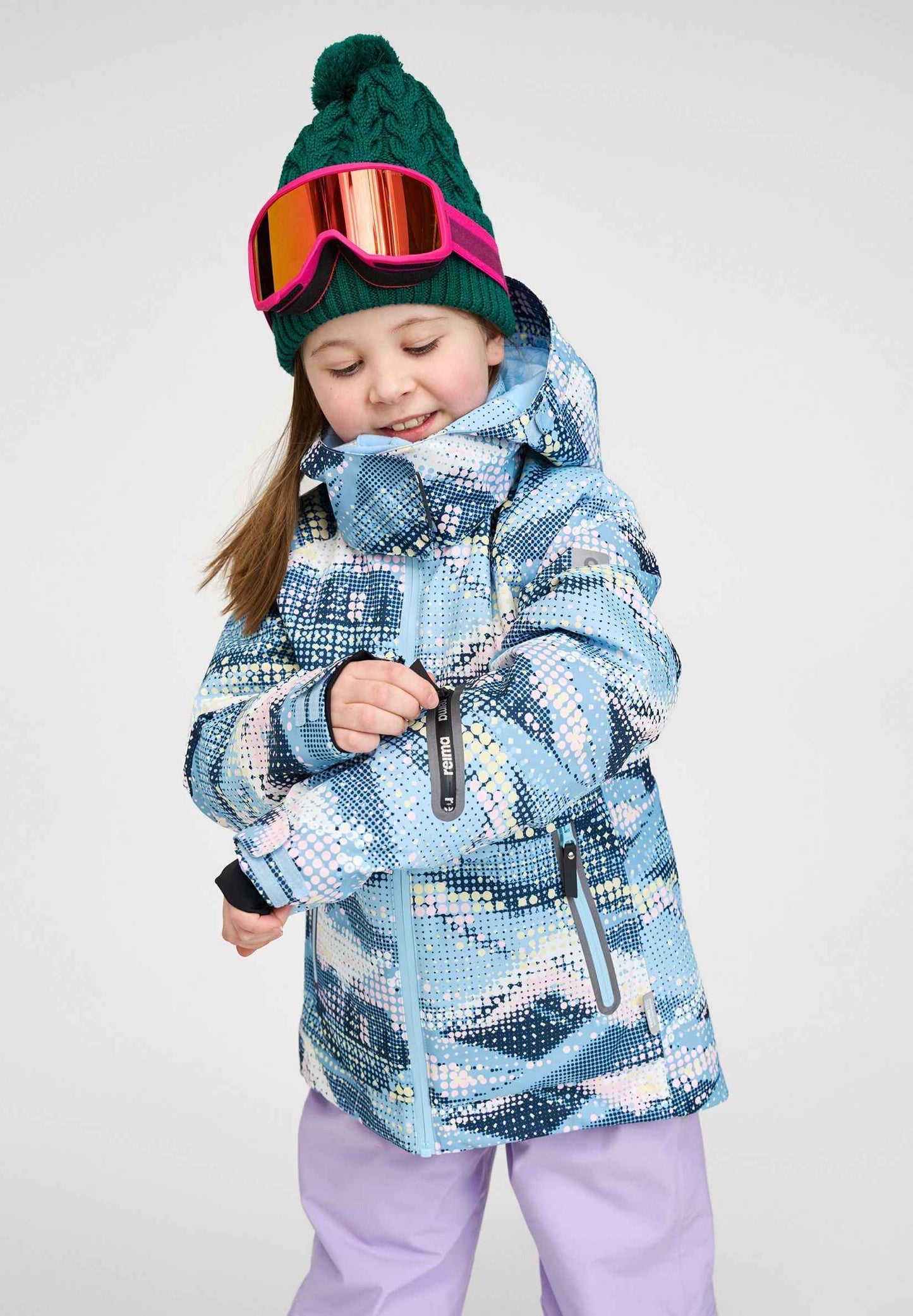Lifestyle portrait of a young girl wearing the frozen blue Reima Posio kids' waterproof ski jacket, adjusting the sleeve pass pocket before a day of winter sports.