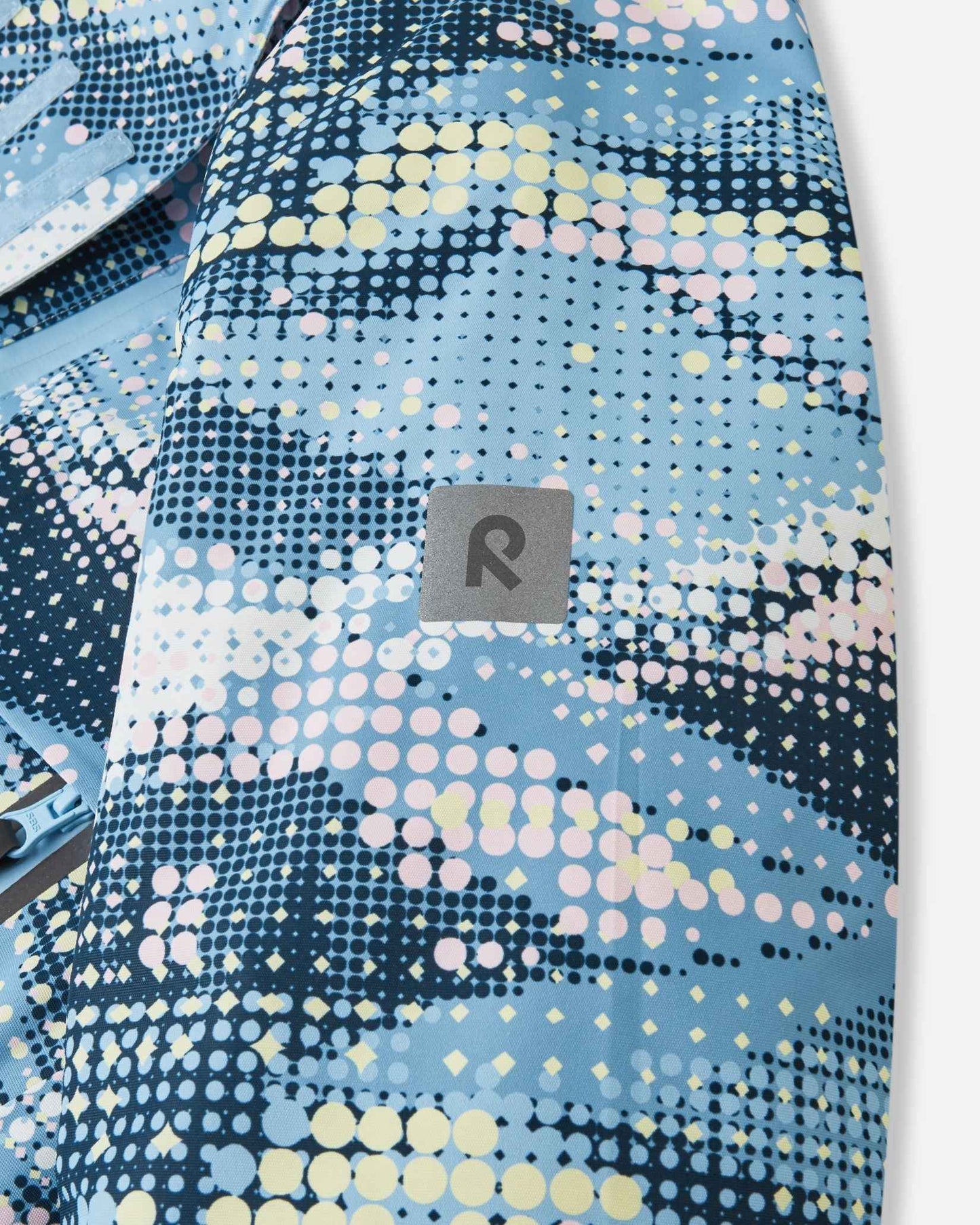 Close-up of the safety-enhancing reflective Reima logo patch on the sleeve of the frozen blue Posio kids' waterproof ski jacket, providing essential visibility in low-light winter conditions.