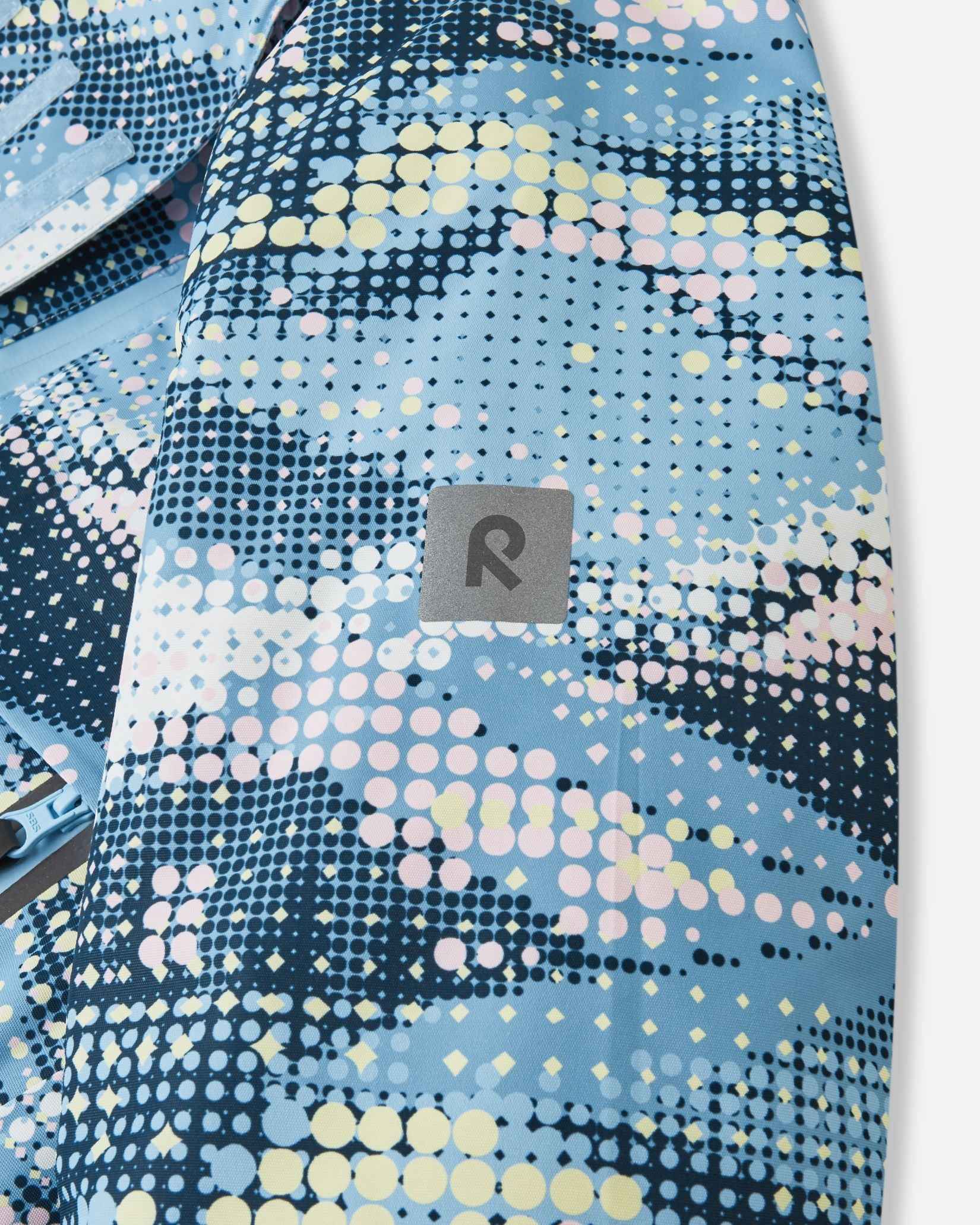Close-up of the safety-enhancing reflective Reima logo patch on the sleeve of the frozen blue Posio kids' waterproof ski jacket, providing essential visibility in low-light winter conditions.