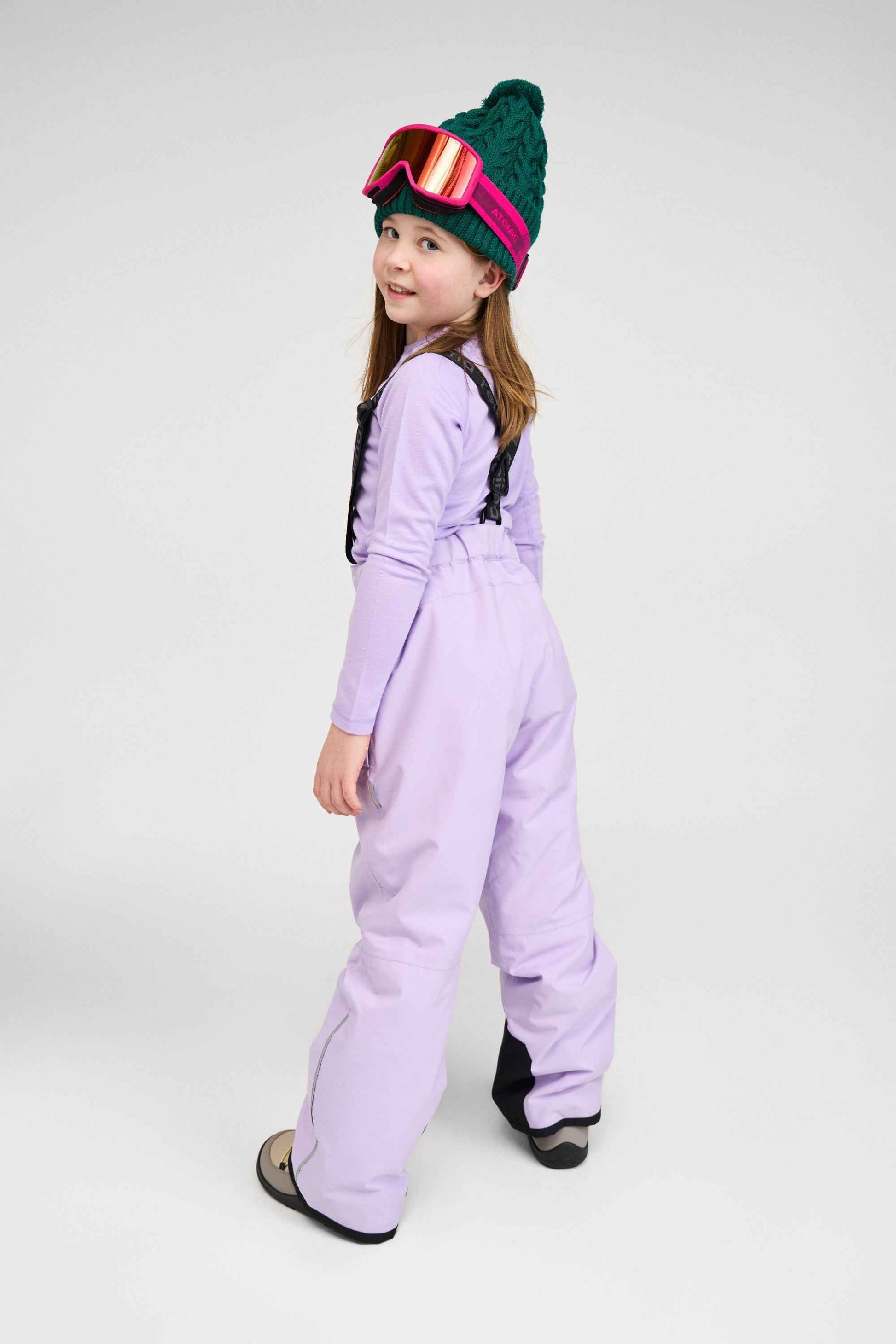 Lifestyle portrait of a young girl from behind wearing the blooming lilac Reima Proxima kids' winter ski pants, a matching long-sleeve base layer, a green knit beanie, and ski goggles. The image highlights the comfortable fit and mobility of the suspenders.