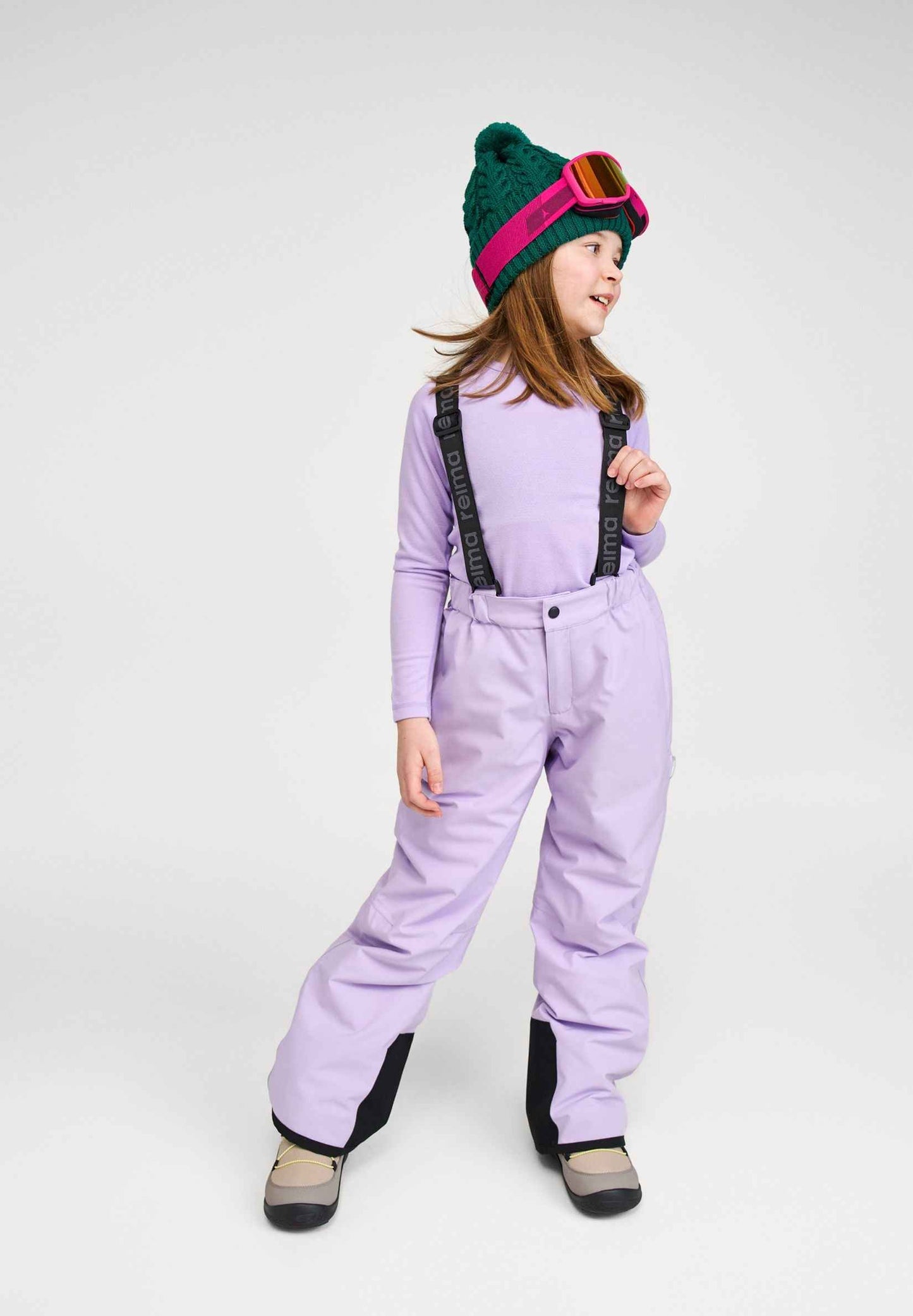 Lifestyle portrait of a young girl smiling while wearing the blooming lilac Reima Proxima kids' winter ski pants, a purple base layer top, green winter beanie, and pink ski goggles, showcasing the practical fit and style for young skiers.