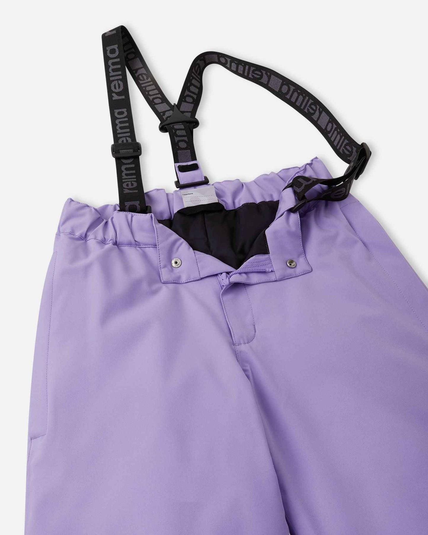 Close-up detail of the front waist on the blooming lilac Reima Proxima kids' ski pants, showing the secure snap buttons, smooth zipper fly, elastic back waistband, and adjustable branded suspenders.