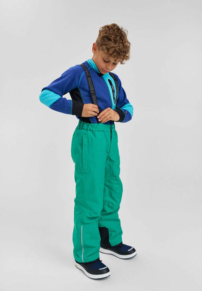 Lifestyle portrait of a young boy wearing the bright green Reima Proxima kids' winter ski pants paired with a blue fleece jacket and winter boots, demonstrating the comfortable fit, mobility, and high visibility for outdoor snow sports.Reima Proxima Ski Pants - Green
