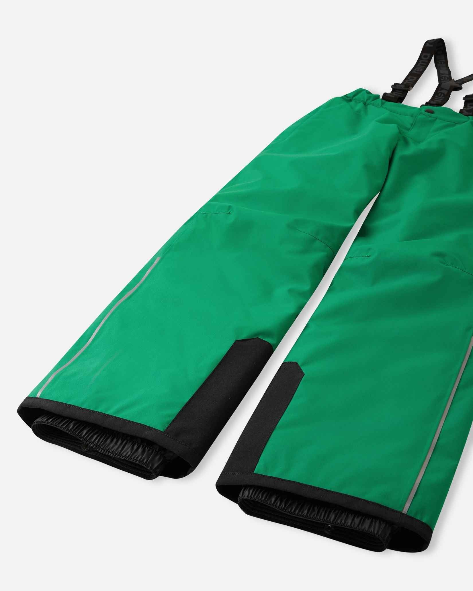 Detail view of the lower legs on the bright green Reima Proxima kids' ski pants, emphasizing the rugged black reinforced hems to protect against ski edges and the inner elastic snow locks designed to securely grip boots and keep powder out.