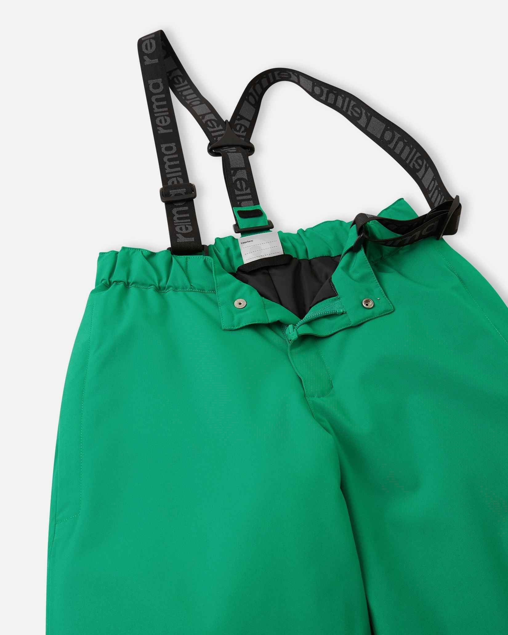 Close-up detail of the front waist on the bright green Reima Proxima kids' ski pants, showing the secure snap buttons, smooth zipper fly, elastic back waistband, and adjustable branded suspenders.