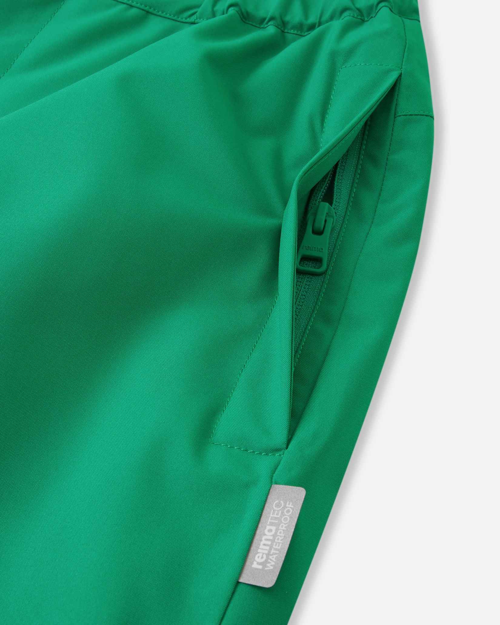 Close-up detail of the hip on the bright green Reima Proxima kids' ski pants, highlighting a secure, low-profile zippered pocket and a Reimatec waterproof quality tag.