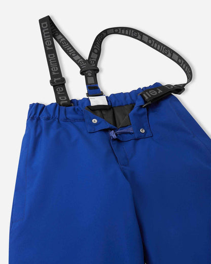 Close-up detail of the front waist on the twilight blue Reima Proxima kids' ski pants, showing the secure snap buttons, smooth zipper fly, elastic back waistband, and adjustable branded suspenders.