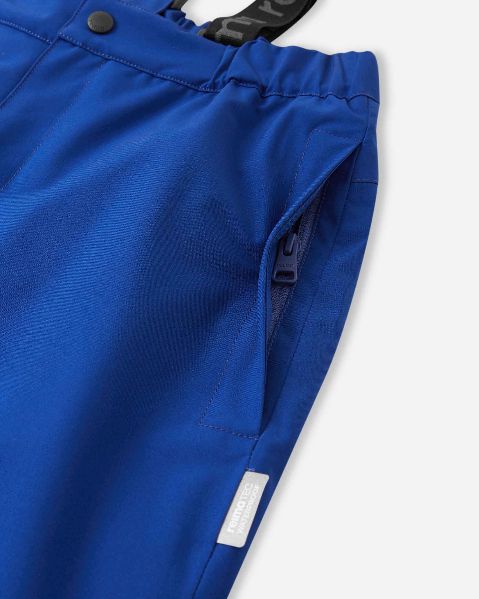 Close-up detail of the hip on the twilight blue Reima Proxima kids' ski pants, highlighting a secure, low-profile zippered pocket and a Reimatec waterproof quality tag.