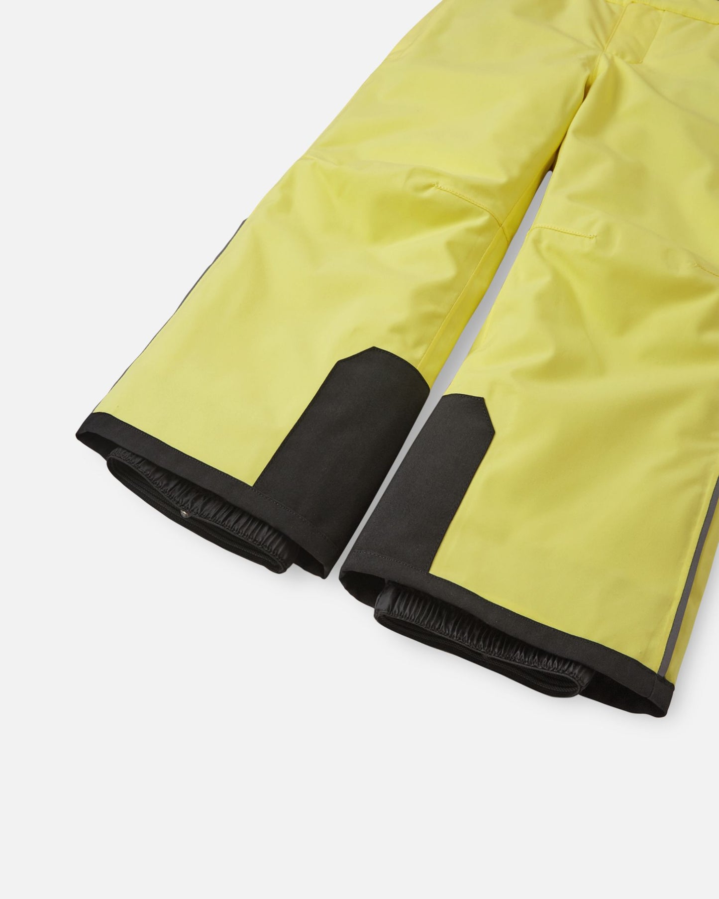 Detail view of the lower legs on the bright yellow Reima Proxima kids' ski pants, emphasizing the rugged black reinforced hems to protect against ski edges and the inner elastic snow locks designed to securely grip boots and keep powder out.