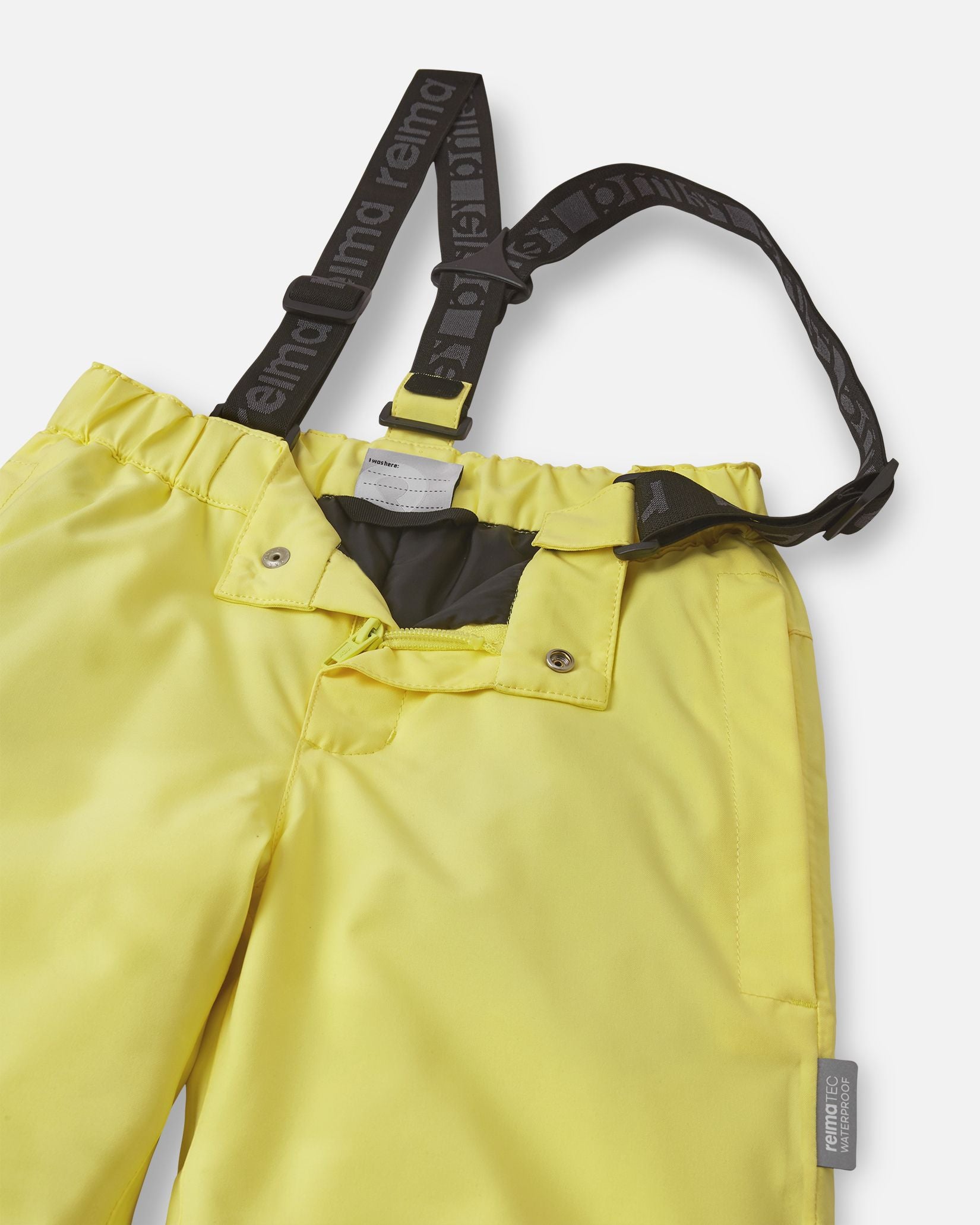 Close-up detail of the front waist on the bright yellow Reima Proxima kids' ski pants, showing the secure snap buttons, smooth zipper fly, elastic back waistband, and adjustable branded suspenders.