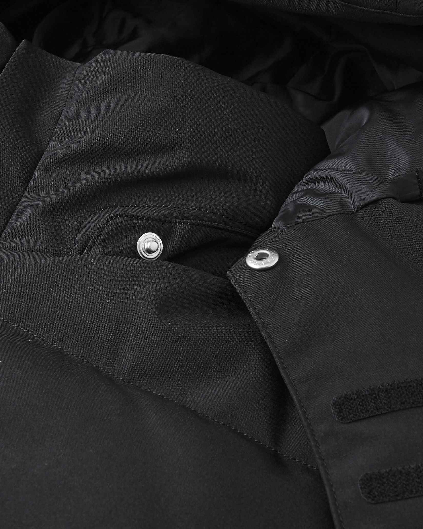 Close-up of the collar area on the Reima Ranua kids' down ski jacket in black, detailing the secure snap buttons used to attach the safe, easily removable winter hood.