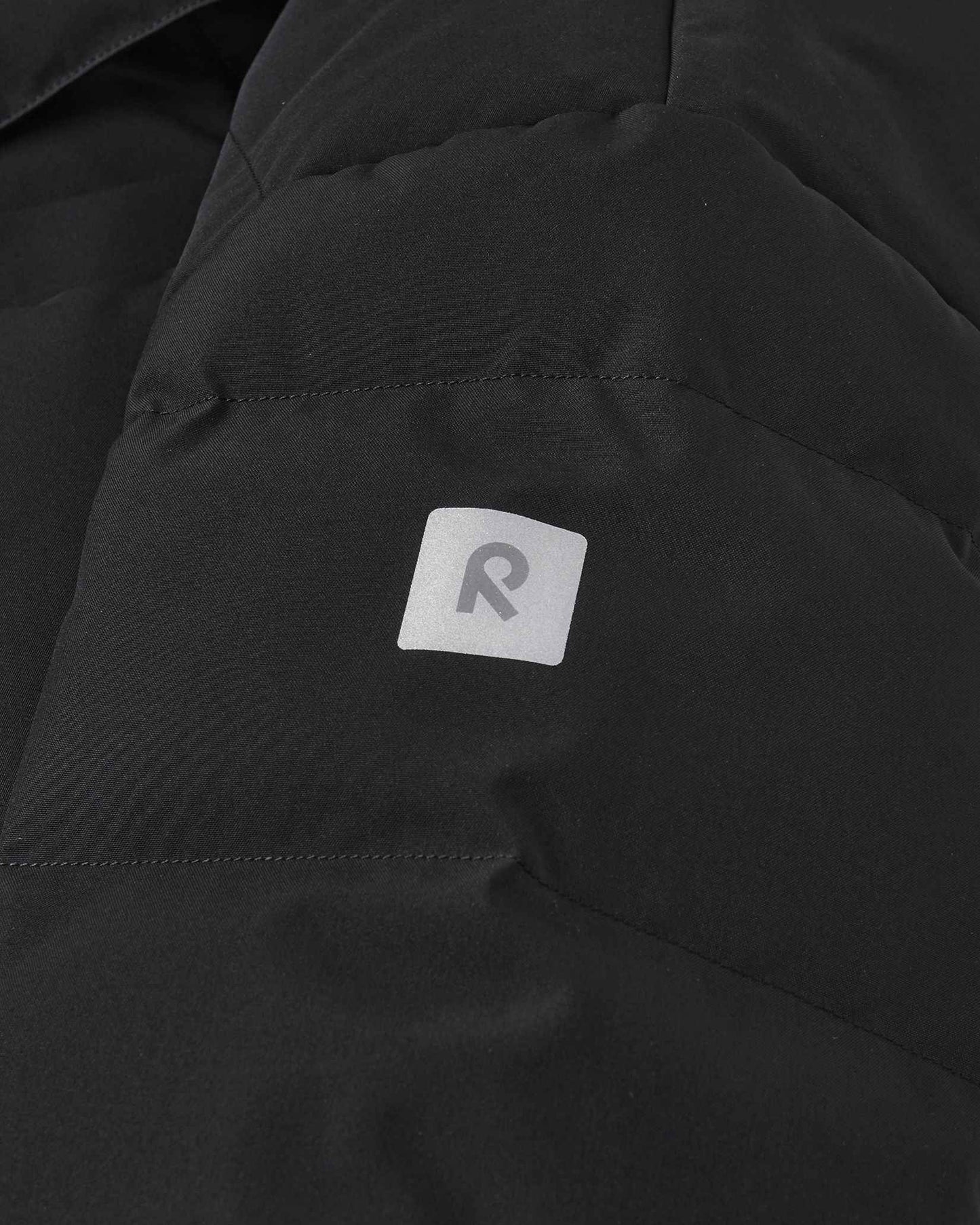 Close-up detail of the black Reima Ranua kids' down ski jacket, highlighting the horizontal down insulation quilting and a square reflective Reima logo patch designed to enhance winter safety and visibility.