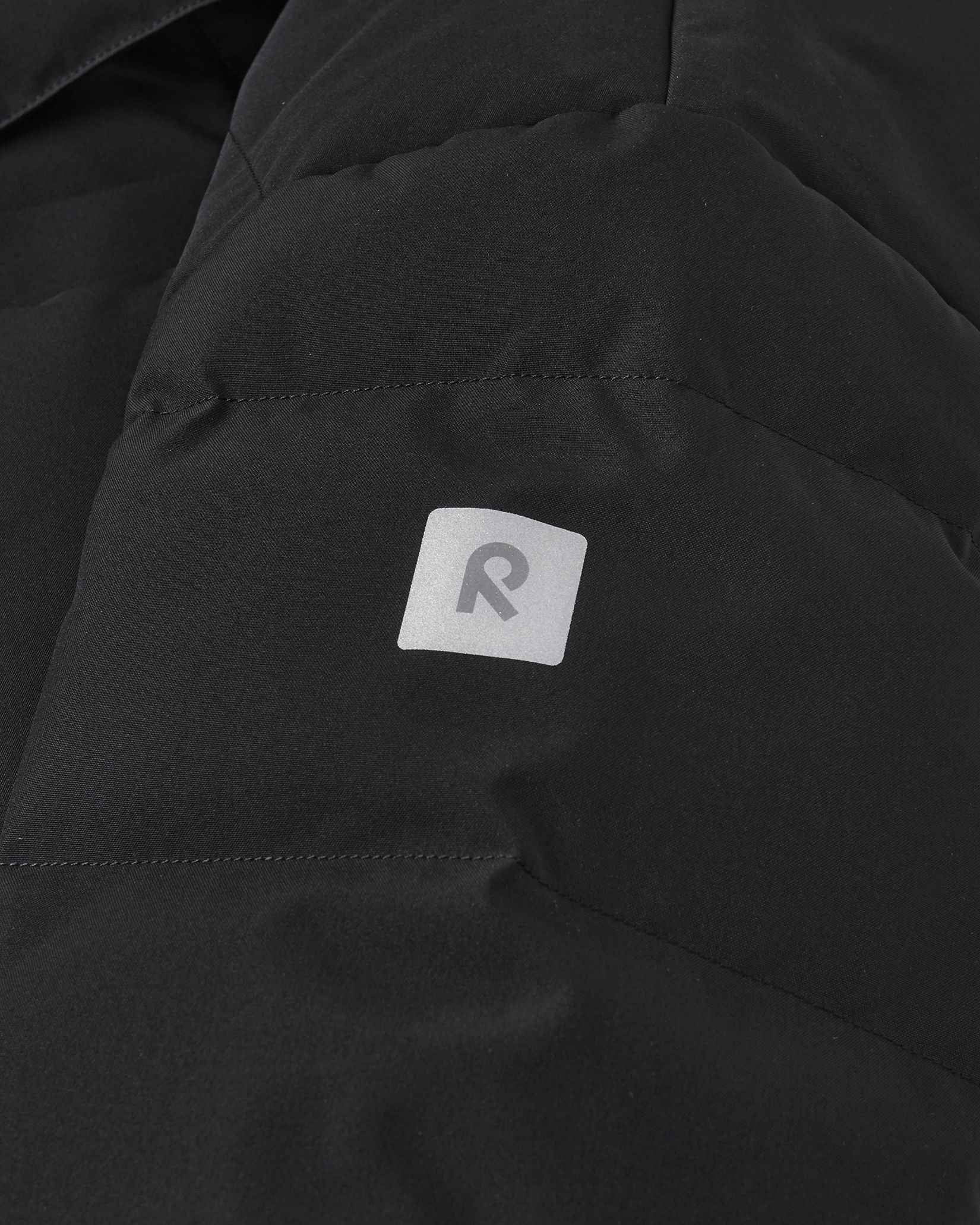 Close-up detail of the black Reima Ranua kids' down ski jacket, highlighting the horizontal down insulation quilting and a square reflective Reima logo patch designed to enhance winter safety and visibility.