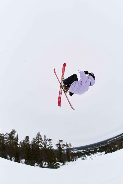 An extreme wide-angle action shot from below of a skier catching massive air during a snow park jump, wearing the blooming lilac Reima Ranua kids' down ski jacket and black ski pants, with bright red skis framed against a stark white winter sky above a snowy forest.