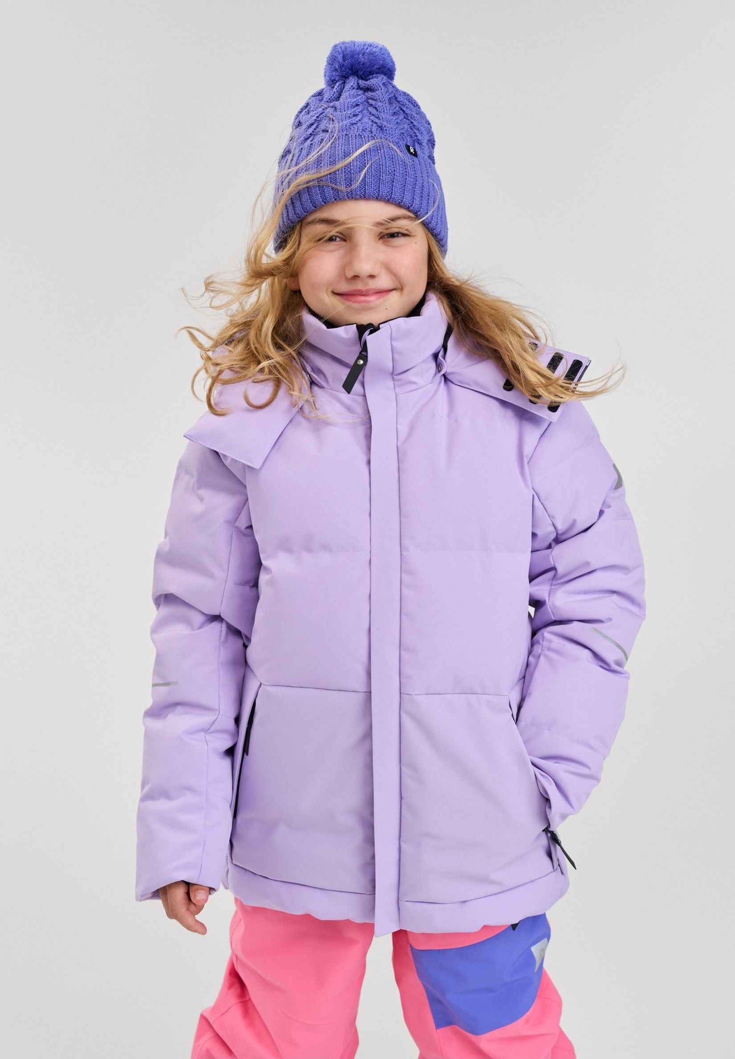 A smiling young girl modeling the blooming lilac Reima Ranua kids' down ski jacket in a studio setting, paired with bright pink and blue snow pants and a purple knit beanie, demonstrating a complete, stylish, and warm winter outdoor outfit.