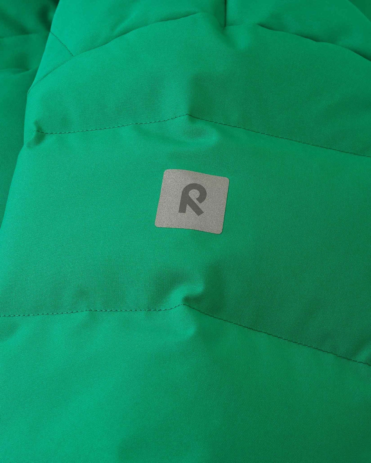 Close-up detail of the green Reima Ranua kids' down ski jacket, highlighting the horizontal down insulation quilting and a square reflective Reima logo patch designed to enhance winter safety and visibility.