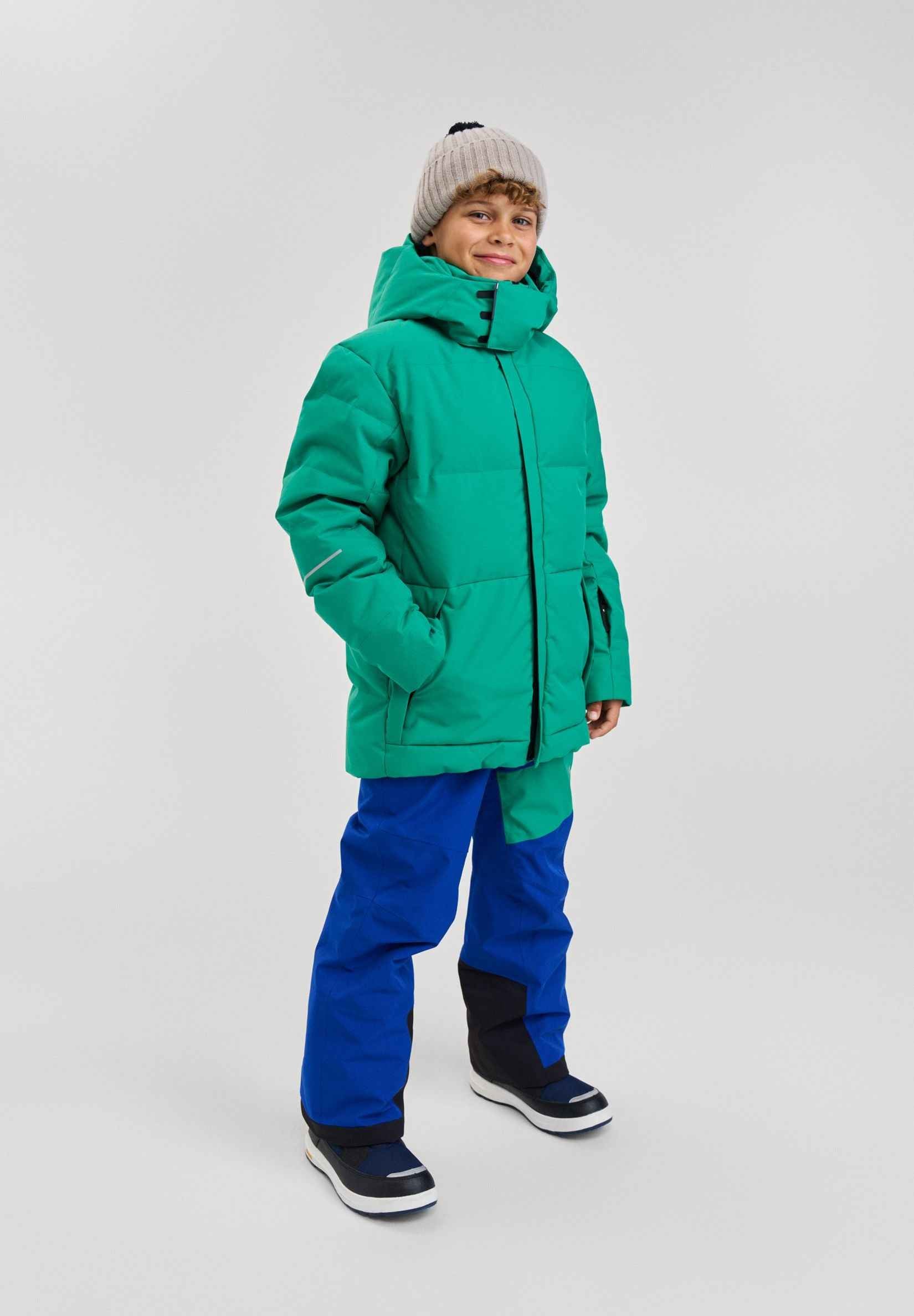 A smiling boy modeling the green Reima Ranua kids' down ski jacket in a studio setting, paired with bright blue snow pants, winter boots, and a knit beanie, demonstrating a complete, stylish, and warm winter outdoor outfit.