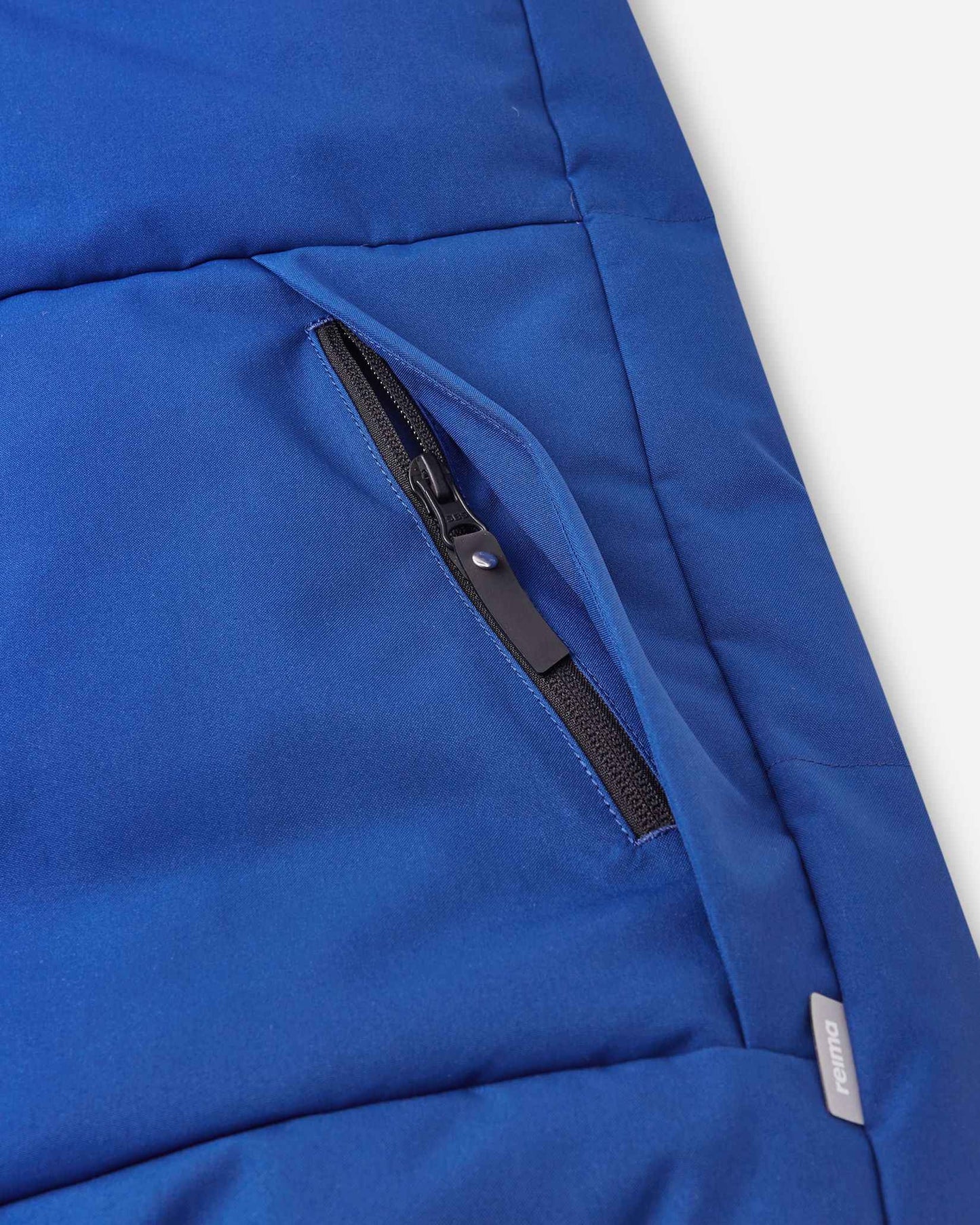 Close-up detail of the secure, hidden front zippered pocket on the quilted twilight blue body of the Reima Ranua kids' down ski jacket, perfect for safely storing small items.