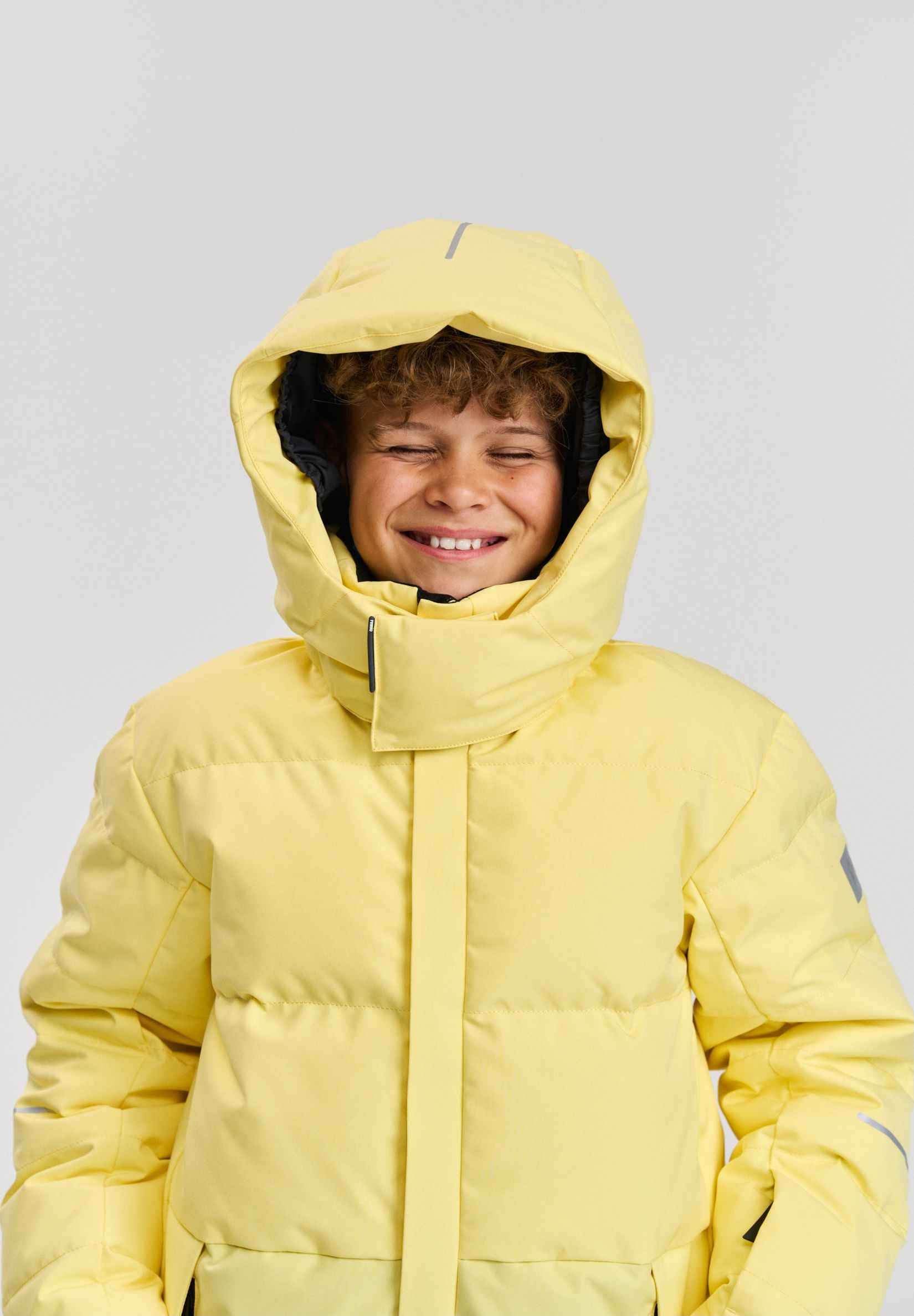 A smiling child wearing the warm Reima Ranua kids' down ski jacket in vibrant yellow with the insulated hood pulled up, demonstrating the comfortable fit and high protective collar designed for winter outdoor activities.