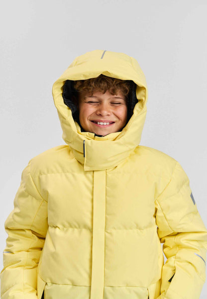 A smiling child wearing the warm Reima Ranua kids' down ski jacket in vibrant yellow with the insulated hood pulled up, demonstrating the comfortable fit and high protective collar designed for winter outdoor activities.