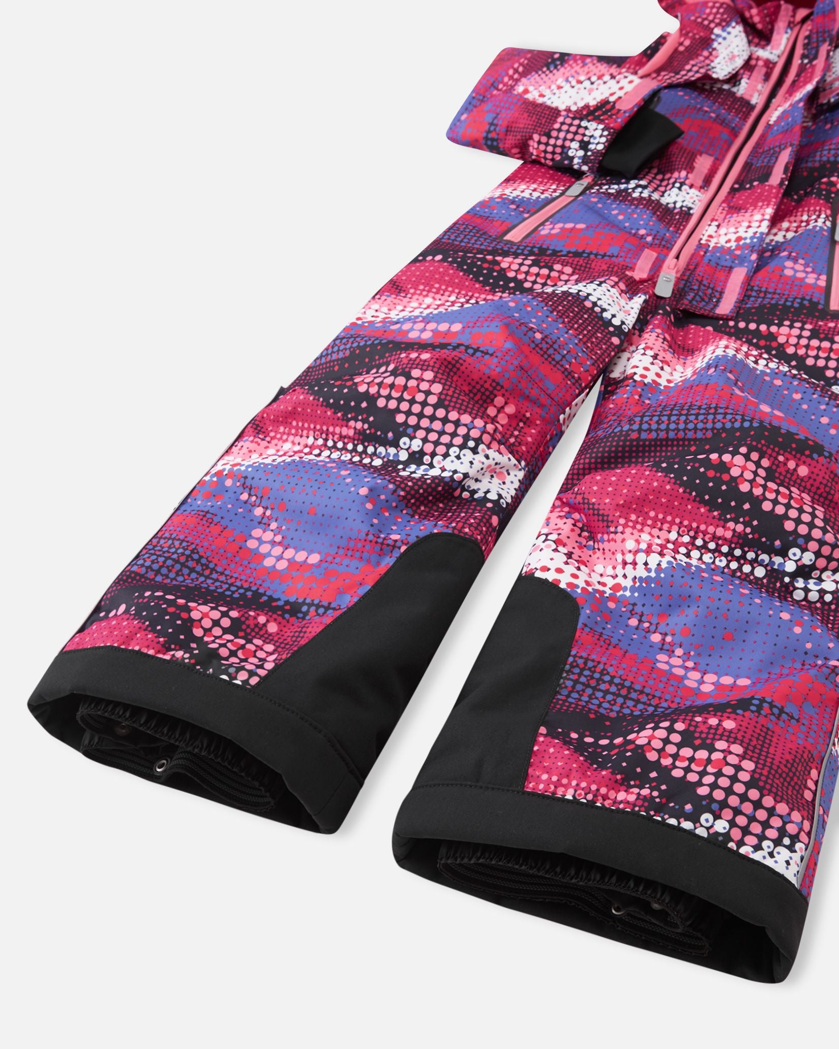 Close-up of the lower legs on the Reima Reach girls' winter ski suit in a rosy berry pattern, showcasing heavy-duty black reinforced kick panels and elastic inner snow gaiters designed to seal tightly over ski boots and block out deep powder.