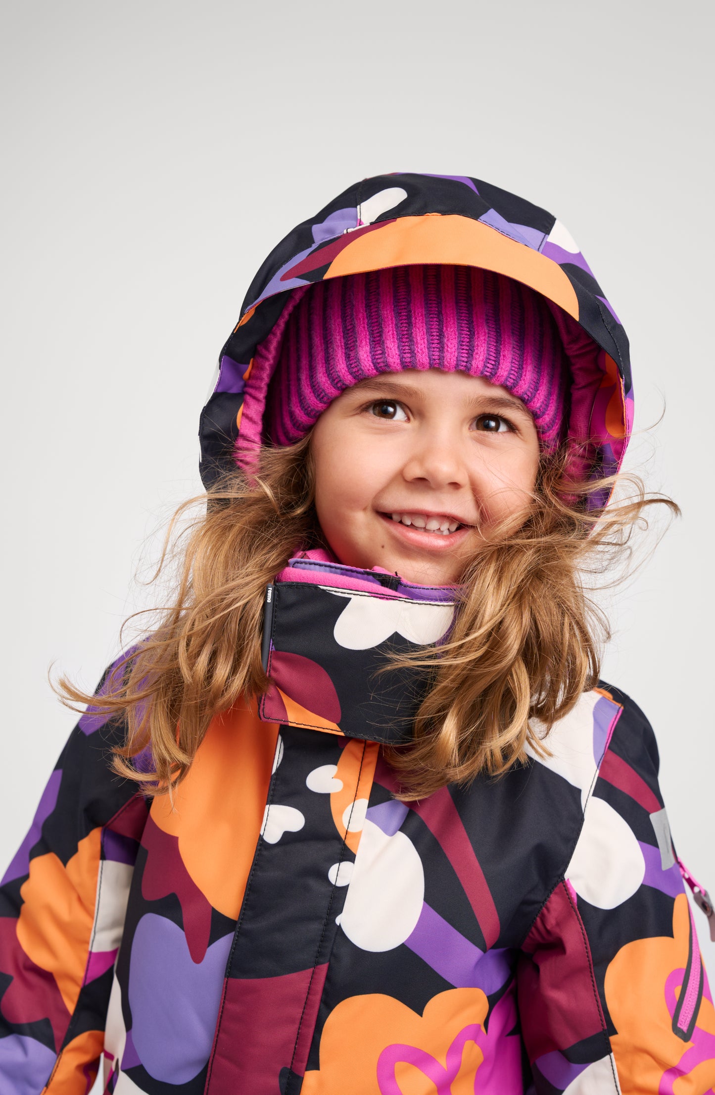 Close-up lifestyle portrait of a smiling child wearing the Reima Reach ski suit in black floral, with the protective storm hood pulled up over a pink winter hat, showing how the high collar and hood combine to provide excellent shelter from winter wind and cold.