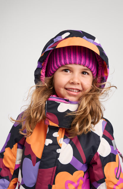 Close-up lifestyle portrait of a smiling child wearing the Reima Reach ski suit in black floral, with the protective storm hood pulled up over a pink winter hat, showing how the high collar and hood combine to provide excellent shelter from winter wind and cold.