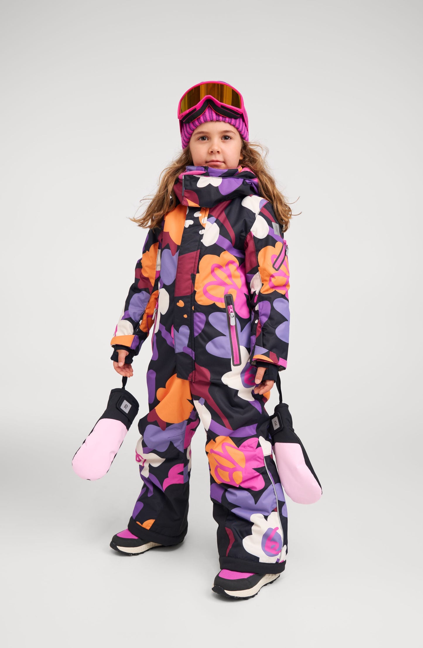 Full-body lifestyle shot of a young girl wearing the Reima Reach kids' ski suit in a vibrant black, purple, orange, and pink floral print. She is geared up for the slopes, holding pink winter mittens and wearing ski goggles, demonstrating the suit's comfortable fit and ease of movement.